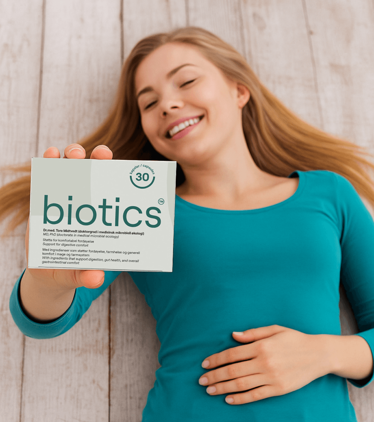 Kvinne ligger med hånden på magen og holder en pakke Biotics postbiotika, 30 kapsler